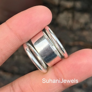 May include: A silver ring with a unique design. The ring features a wide, rectangular center band flanked by two thinner, rounded bands. The ring is held between fingers. The text "SuhaniJewels" is visible.