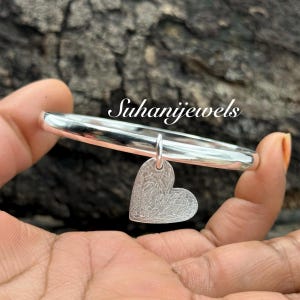 May include: A silver bangle bracelet with a heart-shaped charm. The heart charm has a textured surface. The text "SuhaniJewels" is visible on the image.