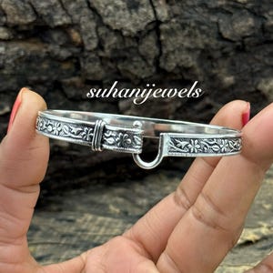 Op de afbeelding: Een zilveren armband met bloemen- en bladerpatronen. De armband heeft een haaksluiting en wordt in een hand gehouden. De tekst "suhanijewels" is zichtbaar op de achtergrond.