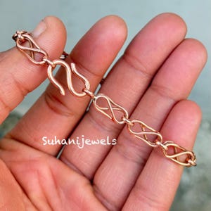 May include: A rose gold bracelet featuring an infinity link design. The bracelet is held in a hand, highlighting the detailed pattern and the S-shaped clasp. The text "Suhanijewels" is visible.