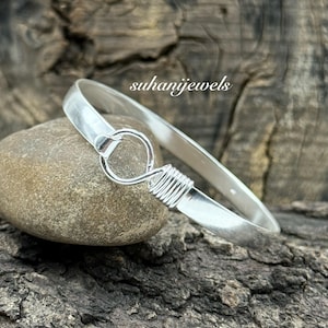 Puede incluir: Una pulsera de plata con cierre de gancho y ojo. La pulsera está sobre una piedra gris y una superficie de madera oscura. El texto "suhanijewels" es visible.