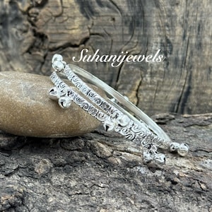 Può includere: Due braccialetti d'argento con intricati motivi floreali e a spirale, adornati da piccole campanelle. I braccialetti poggiano su una pietra liscia marrone chiaro, su uno sfondo di legno strutturato. Il testo "Suhanijewels" è visibile.