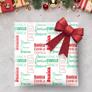 May include: White gift wrap with the name "Danica" in red and green lettering. A large, sparkly red bow adorns the wrapped gift. Festive holiday decorations, including ornaments and wrapped gifts, are in the background.
