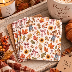 May include: A set of square cork-backed coasters featuring a repeating autumn pattern of scarecrows, sunflowers, wheat stalks, red and orange maple leaves, and pink plaid bows on a white background, arranged in a stacked, overlapping formation.