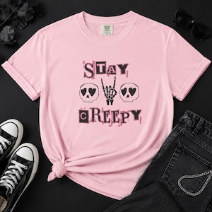 Może przedstawiać: Jasnoróżowy t-shirt z krótkim rękawem z nadrukiem graficznym z napisem STAY CREEPY czarnymi i różowymi literami. Wzór zawiera dwie czaszki z oczami w kształcie serc oraz centralnie umieszczoną dłoń szkieletu wykonującą gest rockowy.