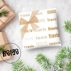 May include: White gift wrap with the name "Travis" printed in gold. A beige ribbon is tied on top. The word "beige" is in a black circle in the lower left corner. Green pine branches are in the background.