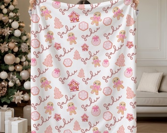 May include: A white blanket featuring a repeating pattern of pink gingerbread men, houses, cookies, and Christmas trees. The name "Sadie" is written in a cursive font throughout the design. The blanket is a cozy home decor item.