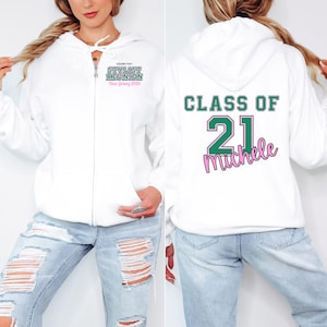 May include: White zip-up hoodie with green and pink text. The front of the hoodie says "STYLIST REUNION New Jersey 2020". The back of the hoodie says "CLASS OF 21 Michele". The hoodie is paired with light blue jeans.