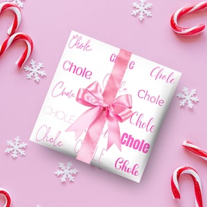 May include: A wrapped gift with white paper printed with the word "Chole" in pink script. A pink satin ribbon and bow adorn the package. Candy canes and snowflake decorations are scattered on a pink background.