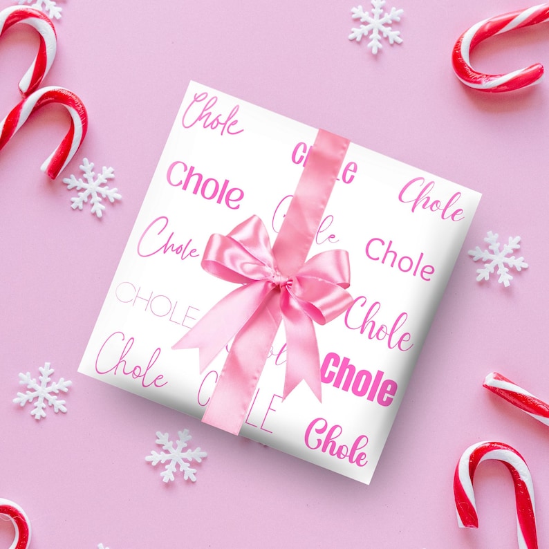 May include: A wrapped gift with white paper printed with the word "Chole" in pink script. A pink satin ribbon and bow adorn the package. Candy canes and snowflake decorations are scattered on a pink background.
