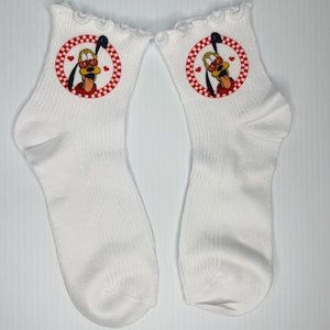 Può includere: Un paio di calzini bianchi con la parte superiore arricciata. Ogni calzino presenta una grafica circolare di Pluto, il cane dei cartoni animati Disney, con un bordo a scacchi rosso e bianco e piccoli cuori rossi.