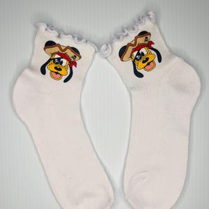 Può includere: Un paio di calzini alla caviglia bianchi con un bordo superiore a balze. Ogni calzino presenta un cane dei cartoni animati con un cappello marrone, una bandana rossa e una faccia gialla. Sono visibili le orecchie e gli occhi neri del cane.