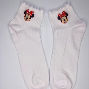 以下が含まれることがあります： ミニーマウスのデザインが施された白いアンクルソックスのペア。各ソックスは、赤いリボンと白い水玉模様のミニーマウスのグラフィックと、フリルのトップが付いています。靴下は柔らかいリブ素材で作られています。