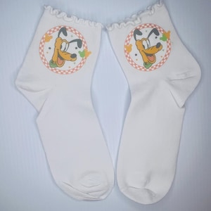 Può includere: Un paio di calzini bianchi con un disegno di un cane dei cartoni animati. Ogni calzino presenta una grafica circolare con la faccia del cane, incorniciata da un bordo a scacchi rosso e arancione. I bordi superiori dei calzini hanno un bordo a balze.