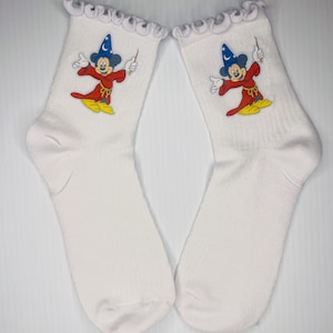 以下が含まれることがあります： フリル付きの白い靴下のペア。各靴下には、魔法使いの帽子とローブを着て杖を持っている漫画のネズミのカラフルなグラフィックが描かれています。マウスは赤、黄、青、黒です。