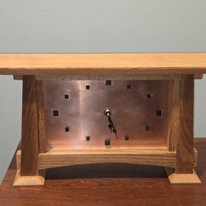May include: A wooden desk clock with a copper face and a light brown wooden frame. The clock face has square cutouts and black clock hands. The clock is rectangular and sits on a wooden surface.