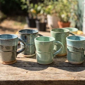 May include: A collection of five ceramic mugs with varied glaze finishes in shades of blue, green, and teal. Each mug features a cylindrical body with a side handle and a distinct horizontal ridge detail. The surfaces show mottled textures.