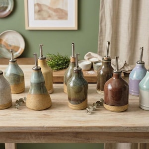 May include: A collection of nine ceramic oil dispensers with various glaze colors including white, blue, yellow, green, brown, and purple. Each bottle features a rounded base, a tapered neck, and a silver-toned metal pour spout on top.