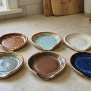 May include: A collection of six round ceramic spoon rests arranged in two rows. Each piece features a unique glaze finish, including speckled brown, light blue, solid off-white, sage green, smooth medium brown, and a deep, vibrant royal blue.
