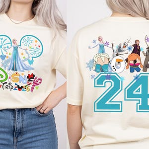 May include: Cream-colored t-shirt featuring a Disney Trip design with Elsa and fireworks. The back of the shirt displays the number 24 in teal, along with various Frozen characters. A fun and colorful design.