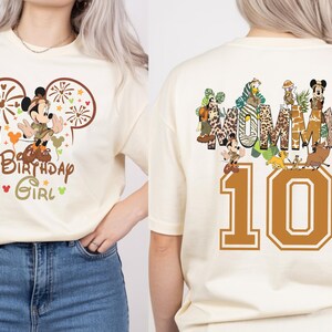 Puede incluir: Camiseta color crema con un dise&ntilde;o de Mickey Mouse, el texto "Birthday Girl" y el n&uacute;mero "10". La parte trasera de la camiseta tiene la palabra "MUMMY" y varios personajes de dibujos animados.