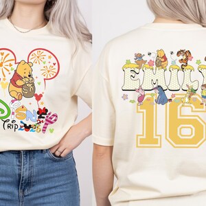 May include: Cream-colored t-shirt featuring a Disney Trip 2023 graphic with Winnie the Pooh. The back of the shirt displays the name "Emily" and the number "16" with Disney characters. The design is in yellow, red, and various colors.