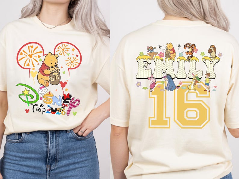 May include: Cream-colored t-shirt featuring a Disney Trip 2023 graphic with Winnie the Pooh. The back of the shirt displays the name "Emily" and the number "16" with Disney characters. The design is in yellow, red, and various colors.