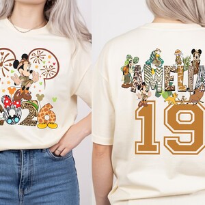 May include: Cream-colored t-shirt featuring a Mickey Mouse graphic with the numbers "2026" and the name "AMELIA" on the back. The design includes animal print patterns and cartoon characters. The number "19" is also on the back.