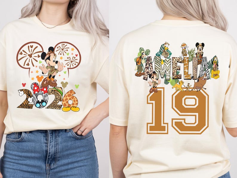 May include: Cream-colored t-shirt featuring a Mickey Mouse graphic with the numbers "2026" and the name "AMELIA" on the back. The design includes animal print patterns and cartoon characters. The number "19" is also on the back.