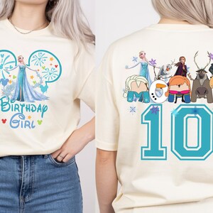 Puede incluir: Camiseta color crema con un gr&aacute;fico frontal de Elsa de Frozen y el texto "Birthday Girl". La parte trasera de la camiseta muestra el n&uacute;mero "10" y otros personajes de Frozen. El dise&ntilde;o es en tonos de azul, turquesa y blanco.
