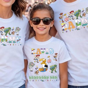 May include: White t-shirts with colorful graphics featuring characters from a popular animated movie. The shirts have the words "MOMMY", "DADDY", and "DAVID" with the words "BIRTHDAY BOY" on the child's shirt. The shirts are worn by three people.
