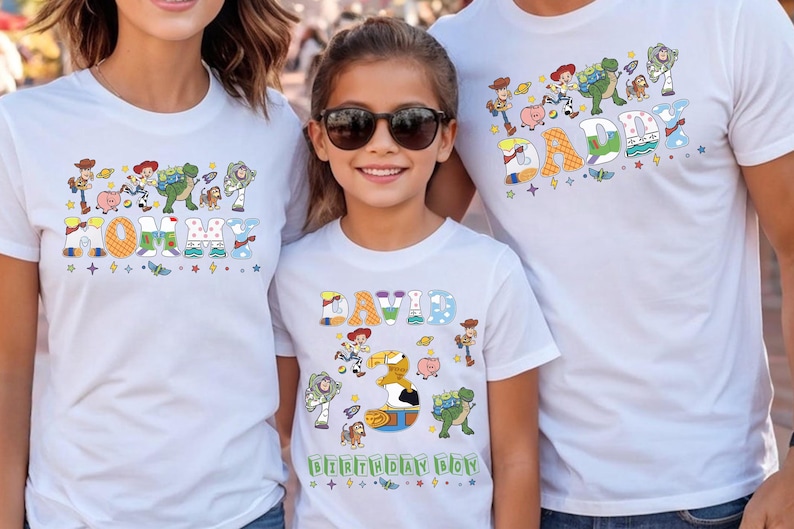 May include: White t-shirts with colorful graphics featuring characters from a popular animated movie. The shirts have the words "MOMMY", "DADDY", and "DAVID" with the words "BIRTHDAY BOY" on the child's shirt. The shirts are worn by three people.