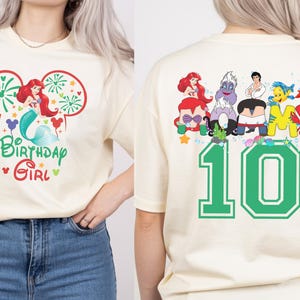 Puede incluir: Camiseta color crema con un gr&aacute;fico colorido de Ariel de La Sirenita, fuegos artificiales y el texto "Birthday Girl". La parte posterior de la camiseta muestra varios personajes y el n&uacute;mero "10" en verde.