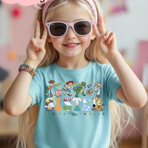 May include: A light blue t-shirt with the word "SISTER" in colorful letters, featuring cartoon characters. The shirt is worn by a child wearing pink sunglasses and a pink bow headband. The image also includes the text "Custom Name & Age".