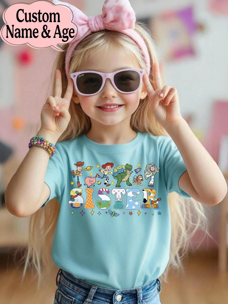 May include: A light blue t-shirt with the word "SISTER" in colorful letters, featuring cartoon characters. The shirt is worn by a child wearing pink sunglasses and a pink bow headband. The image also includes the text "Custom Name & Age".