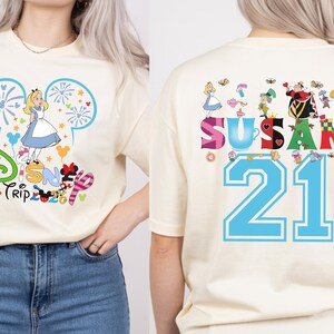 May include: Cream-colored t-shirt with a colorful Disney Trip graphic on the front, featuring Alice in Wonderland. The back of the shirt displays the name "Susan" and the number "21" with Alice in Wonderland characters.