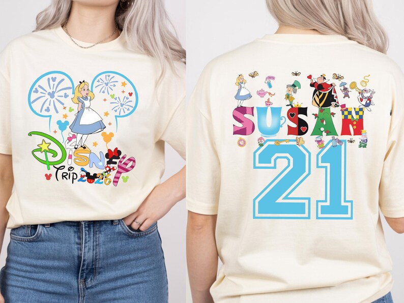 May include: Cream-colored t-shirt with a colorful Disney Trip graphic on the front, featuring Alice in Wonderland. The back of the shirt displays the name "Susan" and the number "21" with Alice in Wonderland characters.