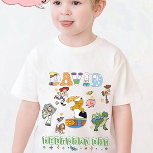 May include: A white t-shirt featuring the name "DAVID" in colorful, playful letters, with the number "3" and various Toy Story characters. The text "BIRTHDAY BOY" is printed at the bottom. The shirt is designed for a child.