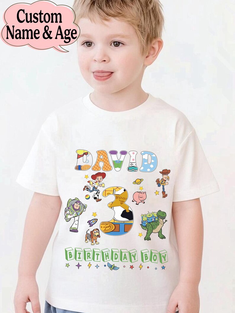 May include: A white t-shirt featuring the name "DAVID" in colorful, playful letters, with the number "3" and various Toy Story characters. The text "BIRTHDAY BOY" is printed at the bottom. The shirt is designed for a child.