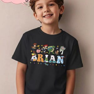 May include: Black t-shirt featuring the name "BRIAN" in colorful, playful letters. The shirt also displays cartoon characters. A pink speech bubble in the upper left corner reads "Custom Name & Age".