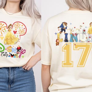 May include: Cream-colored t-shirt featuring a Disney theme. The front displays Belle in a yellow gown within a Mickey Mouse ear outline, with "Disney Trip 2023" text. The back shows the name "LINDA" with Disney characters and the number "17".