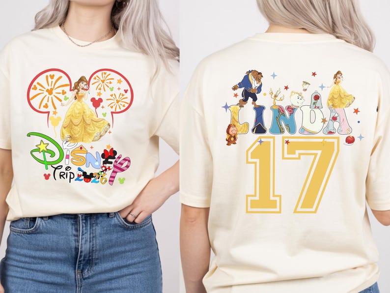 May include: Cream-colored t-shirt featuring a Disney theme. The front displays Belle in a yellow gown within a Mickey Mouse ear outline, with "Disney Trip 2023" text. The back shows the name "LINDA" with Disney characters and the number "17".