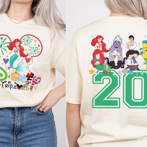 May include: Cream-colored t-shirt featuring a Disney Trip design. The front graphic includes Ariel, fireworks, and the Disney logo. The back displays a green '20' and various Little Mermaid characters.