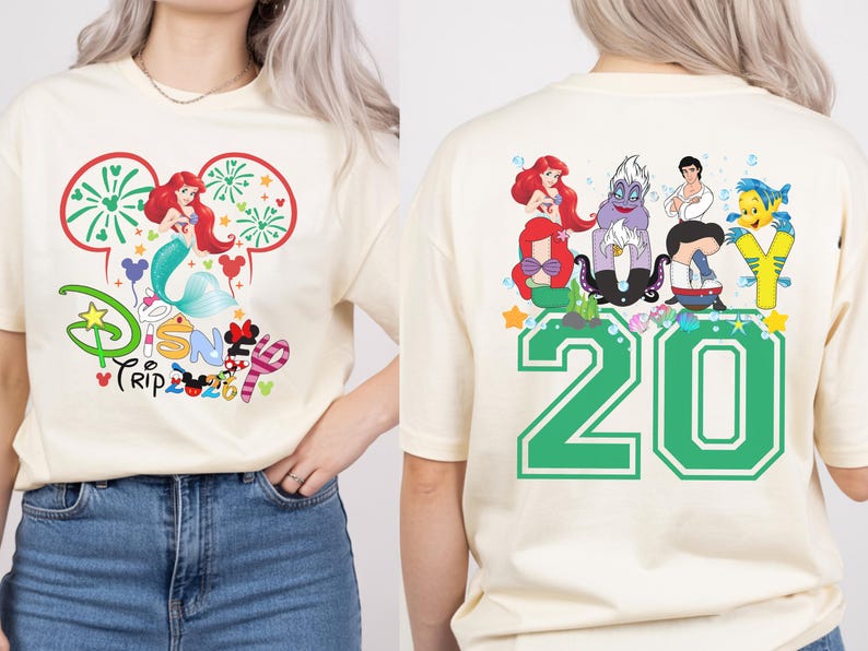 May include: Cream-colored t-shirt featuring a Disney Trip design. The front graphic includes Ariel, fireworks, and the Disney logo. The back displays a green '20' and various Little Mermaid characters.