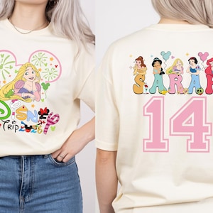 May include: Cream-colored t-shirt featuring a Disney Trip graphic with Rapunzel. The back of the shirt displays the name "SARAH" above the number "14" with Disney princess illustrations.