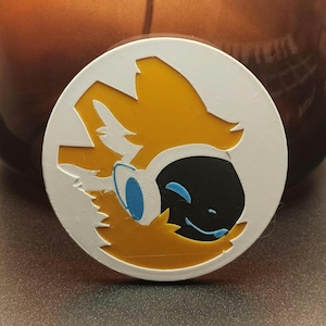 May include: A round, white and yellow graphic featuring a cartoon fox-like character with blue eyes and a black face. The design is set against a blurred background.