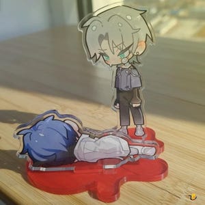 Op de afbeelding: Twee anime-stijl acrylfiguren. Eén figuur staat, met zilver haar, een grijze jas en een zwarte broek. De andere figuur ligt met blauw haar en een wit shirt. Beide figuren staan op een rode basis.