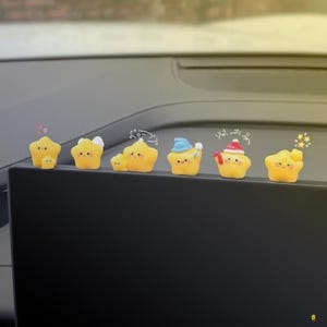 May include: A collection of yellow star-shaped figurines with various accessories, including hats and bows. The figurines are arranged on a black surface, with a blurred background. The stars have smiling faces and are decorated with small details.