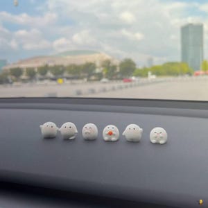 Puede incluir: Una fila de seis pequeñas figuras blancas y redondeadas con rasgos faciales simples y expresiones variadas. Una sostiene una pequeña esfera naranja. Están dispuestas sobre una superficie gris oscuro, con un paisaje urbano borroso al fondo.
