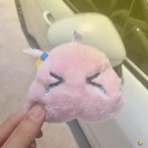 May include: A plush pink character with embroidered black eyes and blue teardrops. The character has a small loop at the top and a small tail. The character is held by a hand.